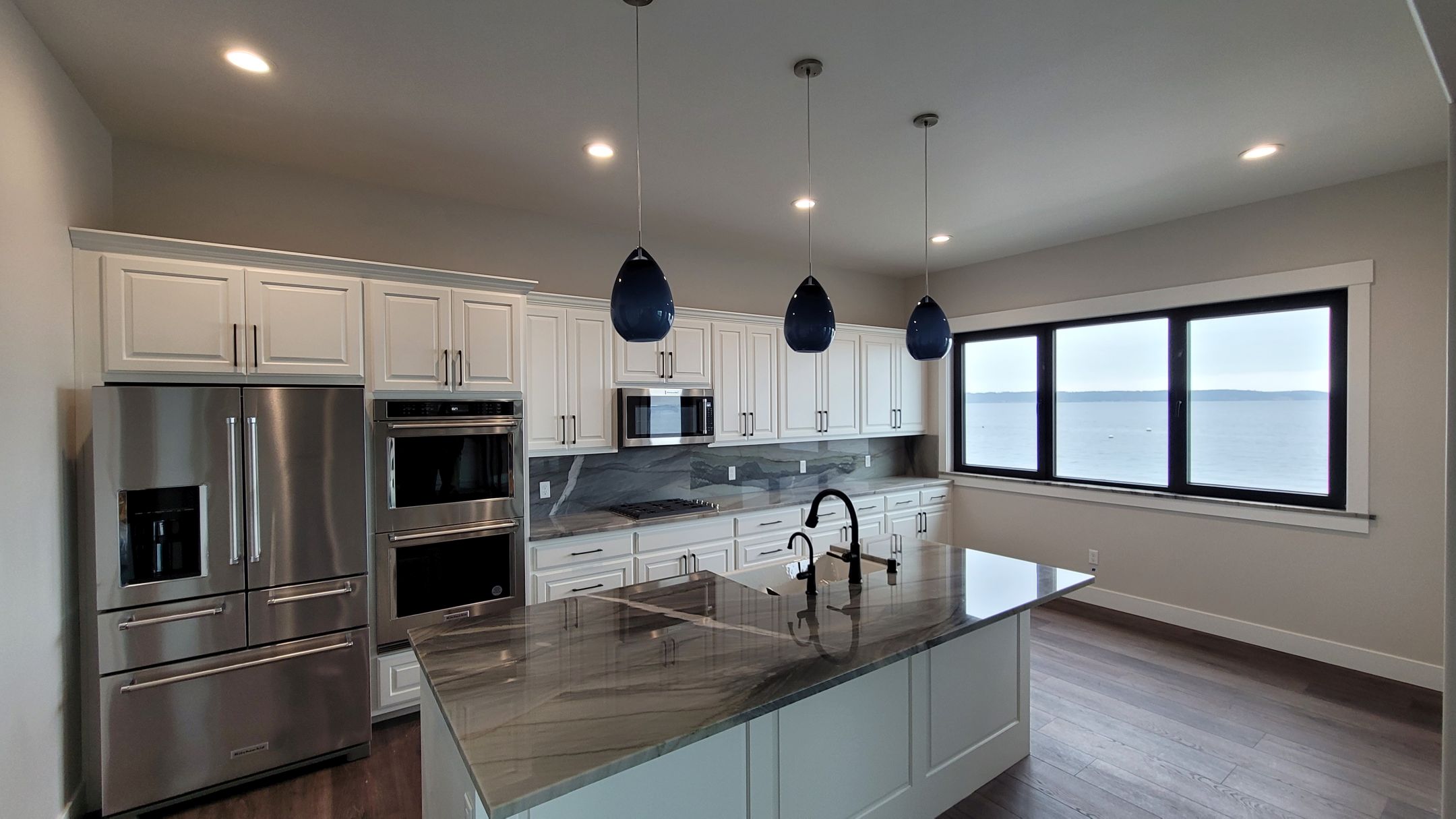 A photo of a modern kitchen with white cabinets, a large island with marble countertops, stainless steel appliances, and a window with a view of a lake.